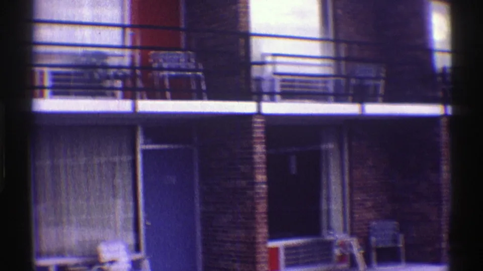 1969 Nebraska motel exterior with two levels of brick units, balcony rails, doors, curtains, and outdoor chairs in home-movie framing.