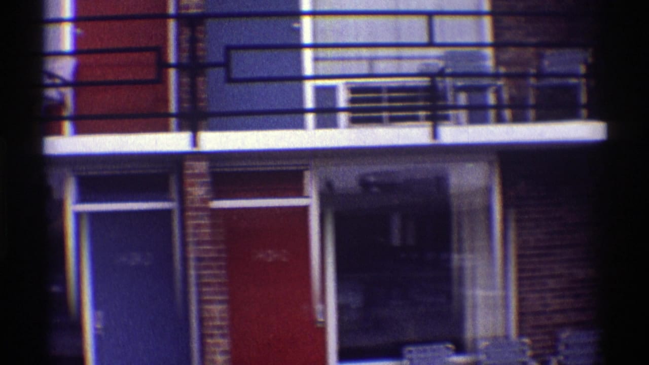 1969 Nebraska home-movie frame showing a brick apartment building with black balcony railings, red and blue doors, and patio chairs.