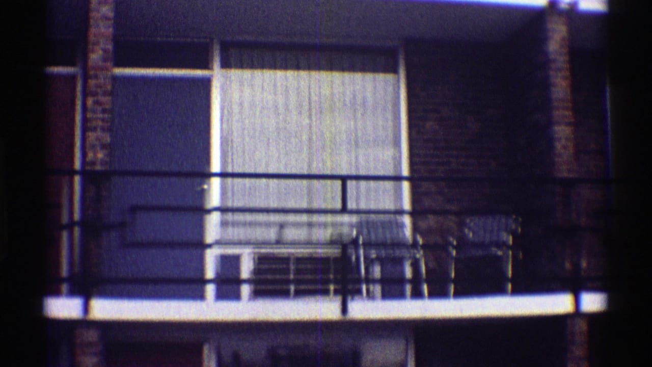 Apartment balcony with black railings, curtained window, and chairs outside.