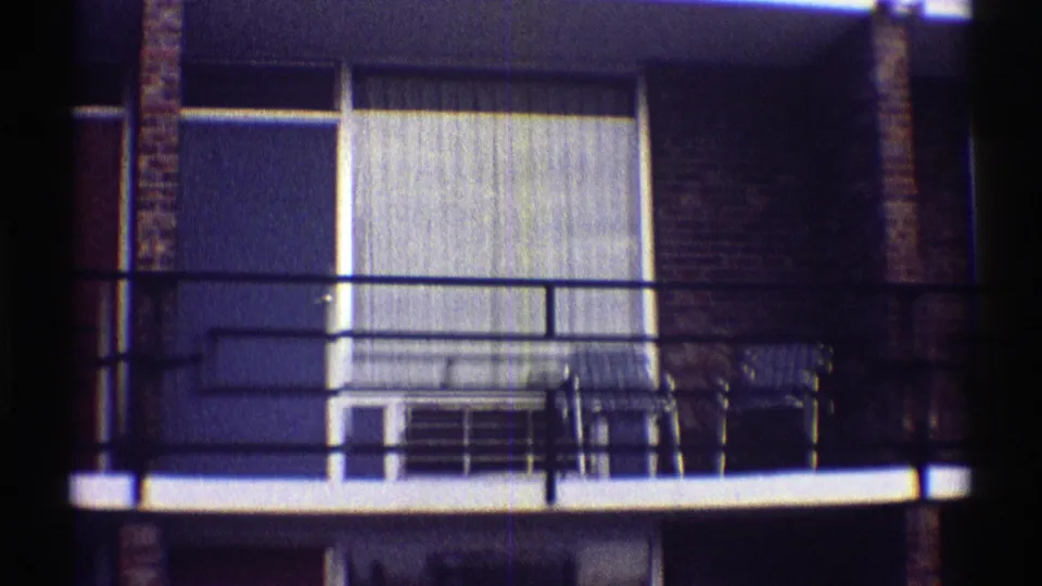 1969 Nebraska apartment balcony with dark metal railings, a large curtained window, and simple outdoor chairs visible on the landing.