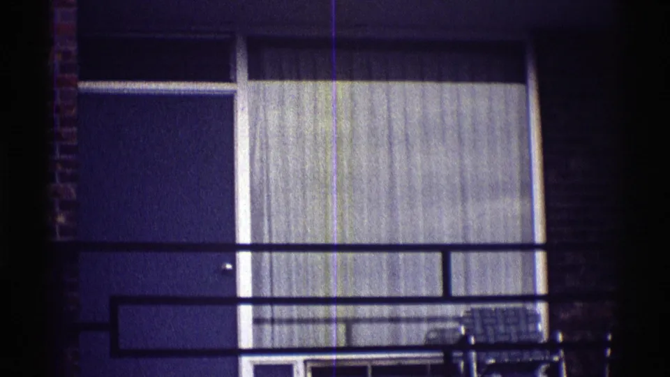 1969 Nebraska residential exterior showing a lower balcony, black metal railing, broad curtained window, and a chair near the building.