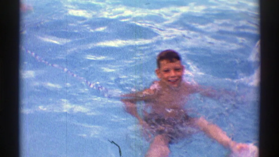 1969 Nebraska home-movie frame with a child visible in an outdoor swimming pool, facing the camera amid bright blue water.
