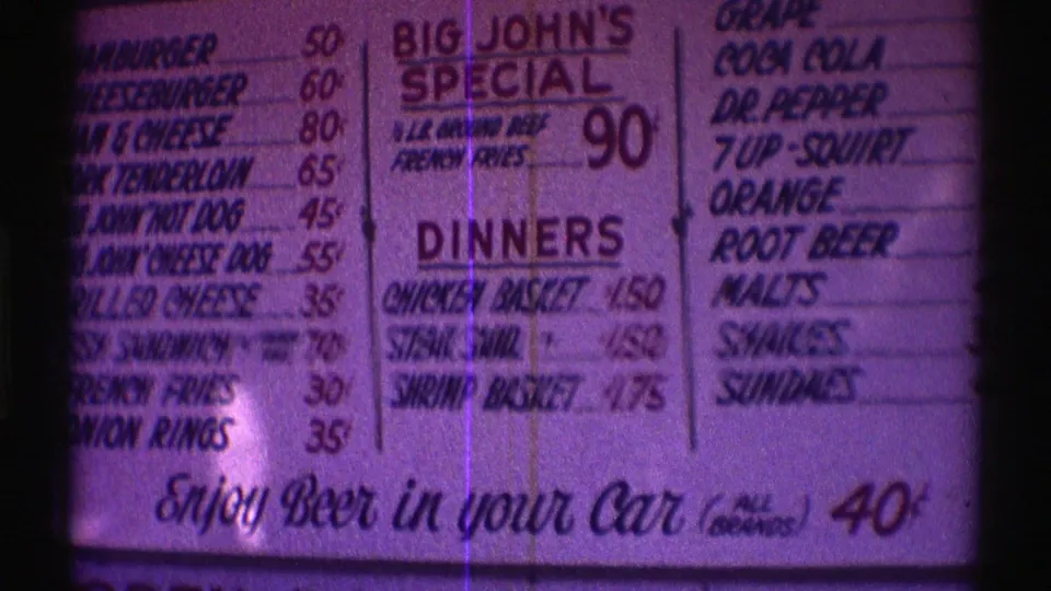 Purple-tinted 8mm frame showing the Nebraska fast food menu with burgers, drinks, dinners, and the beer-in-car line.