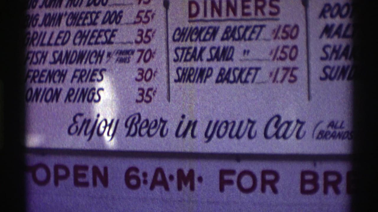 Nebraska 1969 8mm frame showing a close-up diner menu with low food prices and the phrase “Enjoy Beer in your Car.”