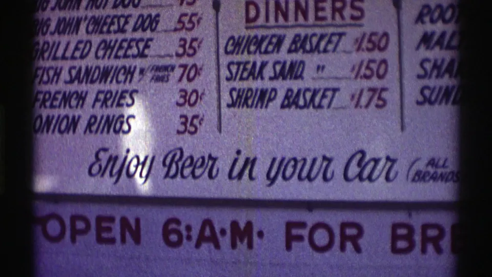 Nebraska 1969 8mm frame showing a close-up diner menu with low food prices and the phrase “Enjoy Beer in your Car.”