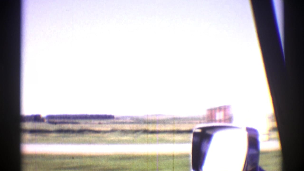Nebraska 1969 home-movie frame looking from a vehicle across open plains, roadside grass, and a car side mirror.