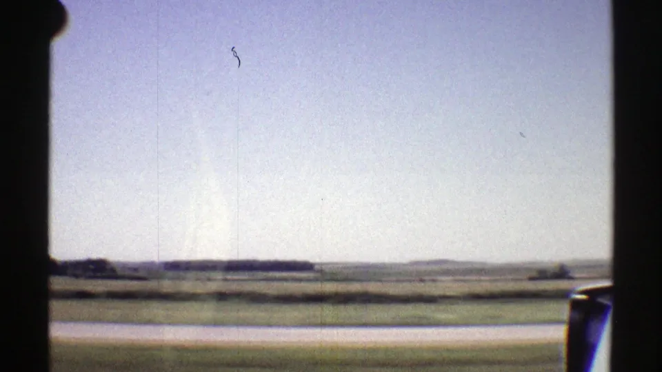 Nebraska 1969 home-movie frame with the car window edge, side mirror, open fields, and a low rural horizon.