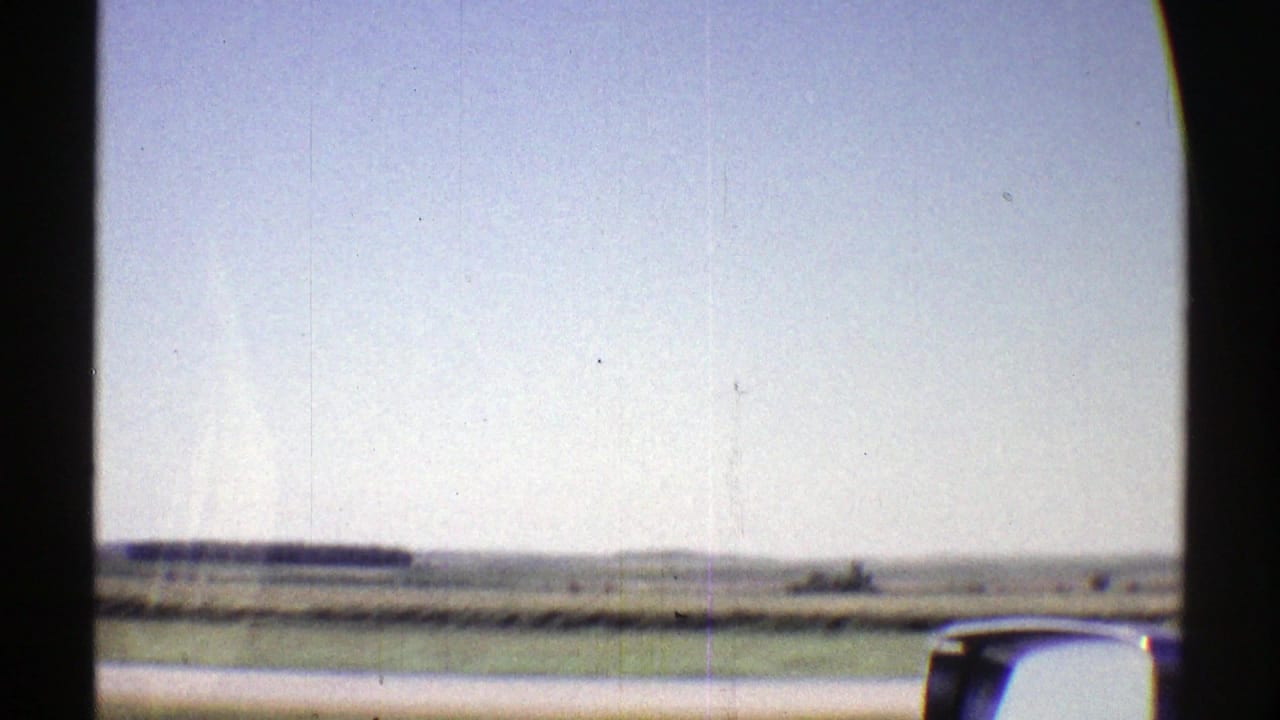 1969 Nebraska home-movie view from a moving car across flat rural fields under a pale sky, with the car window and side mirror visible.