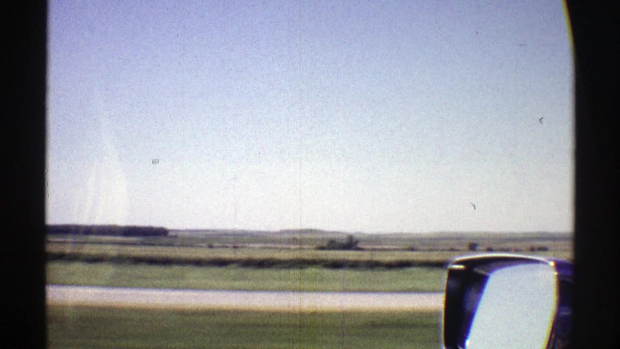 Moving-car 8mm view of Nebraska plains with low fields, distant horizon, pale blue sky, and the vehicle side mirror at right.