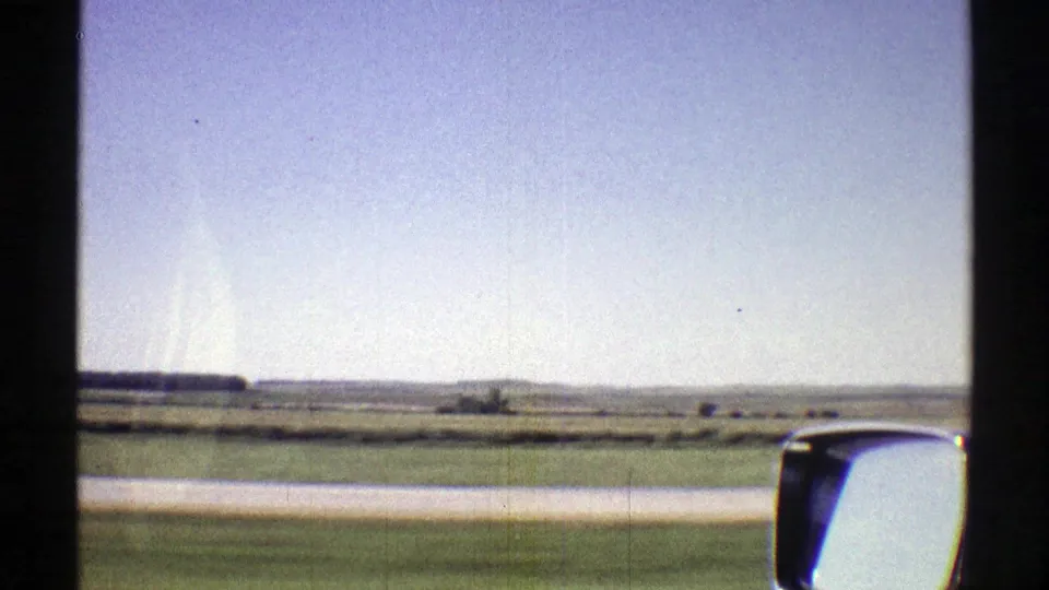 1969 Nebraska road-trip frame showing flat fields, distant dark trees, road shoulder, window frame, and a side mirror in 8mm color.