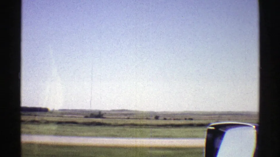 Nebraska plains from a 1969 home movie, filmed through a moving car window with open fields, low horizon, and side mirror in view.