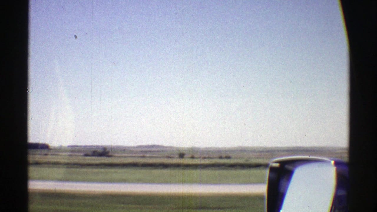 1969 Nebraska 8mm frame from a moving car, showing flat fields, distant low hills, blue sky, and a side mirror.