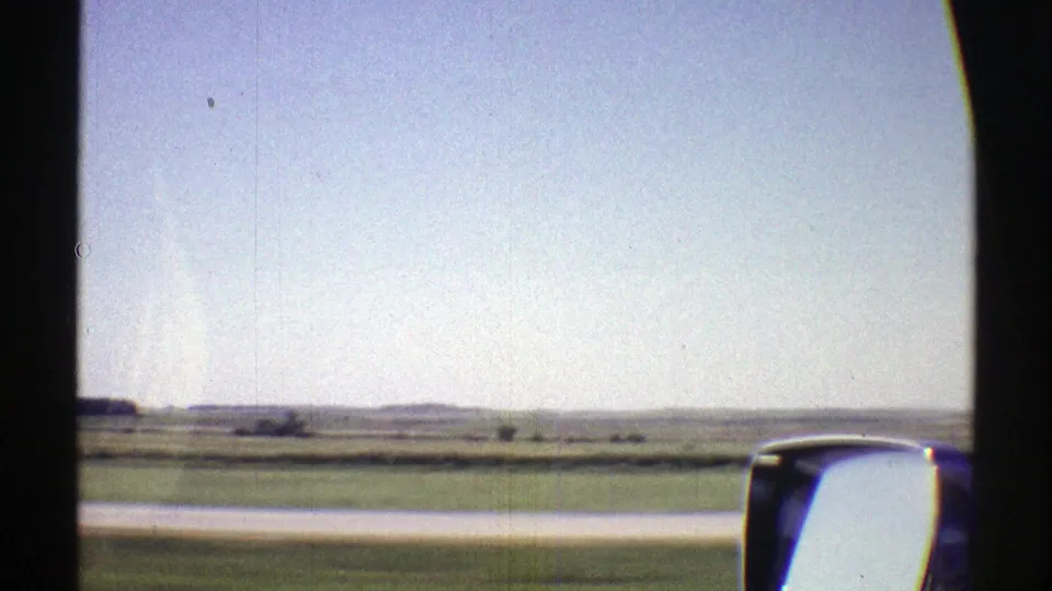 1969 Nebraska 8mm frame from a moving car, showing flat fields, distant low hills, blue sky, and a side mirror.