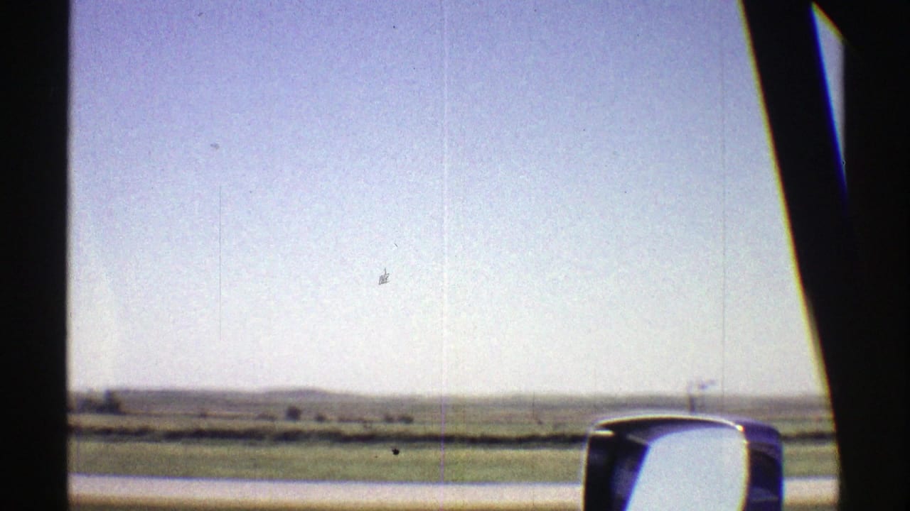 1969 Nebraska road view from inside a moving vehicle, with open fields, low horizon, and side mirror visible.