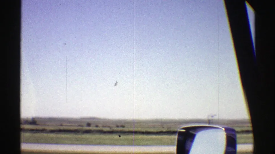 1969 Nebraska road view from inside a moving vehicle, with open fields, low horizon, and side mirror visible.