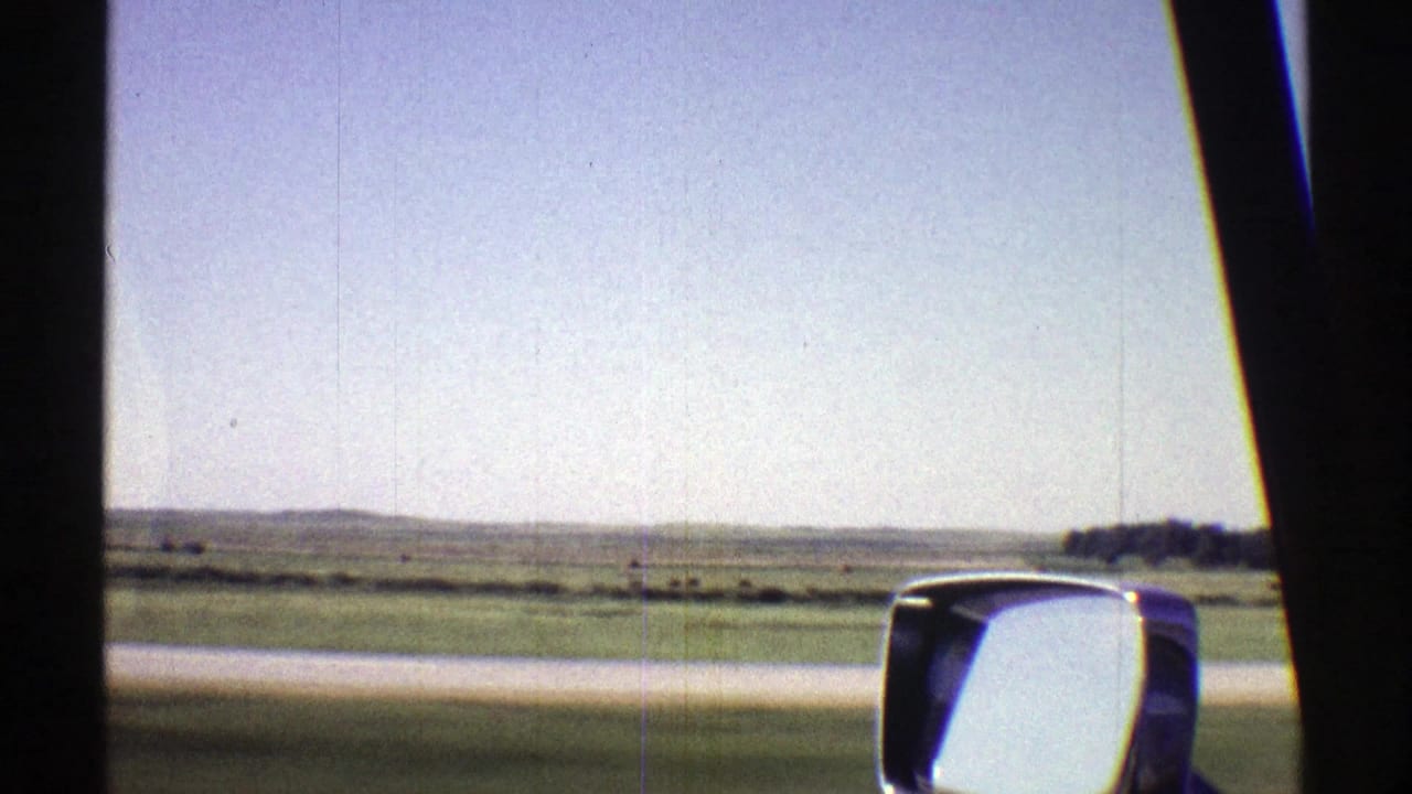 View through a car window of green fields, pale road edge, blue sky, and a chrome side mirror.