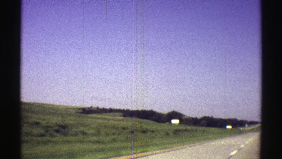 1969 Nebraska moving-car frame showing a paved road beside green fields, blue sky, and small white objects in the distance.