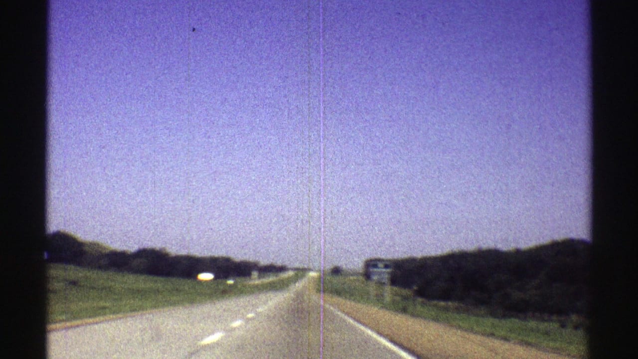 1969 Nebraska road-trip frame with a highway receding ahead, open sky, green roadside slopes, and small signs in the distance.