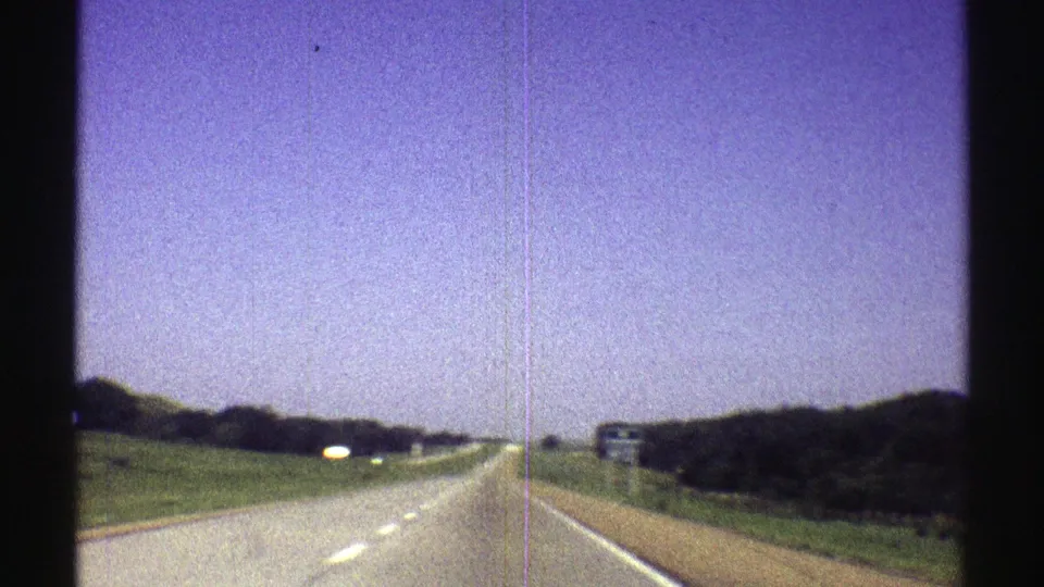 1969 Nebraska road-trip frame with a highway receding ahead, open sky, green roadside slopes, and small signs in the distance.