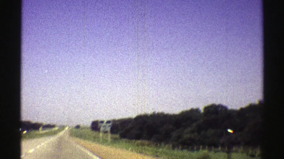 1969 Nebraska 8mm frame showing roadside trees, highway pavement, green margins, and a slightly blurred moving-vehicle perspective.