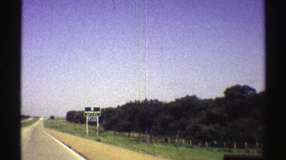 1969 Nebraska frame with a roadside sign, tree line, grassy shoulder, and highway edge captured in 8mm home-movie style.
