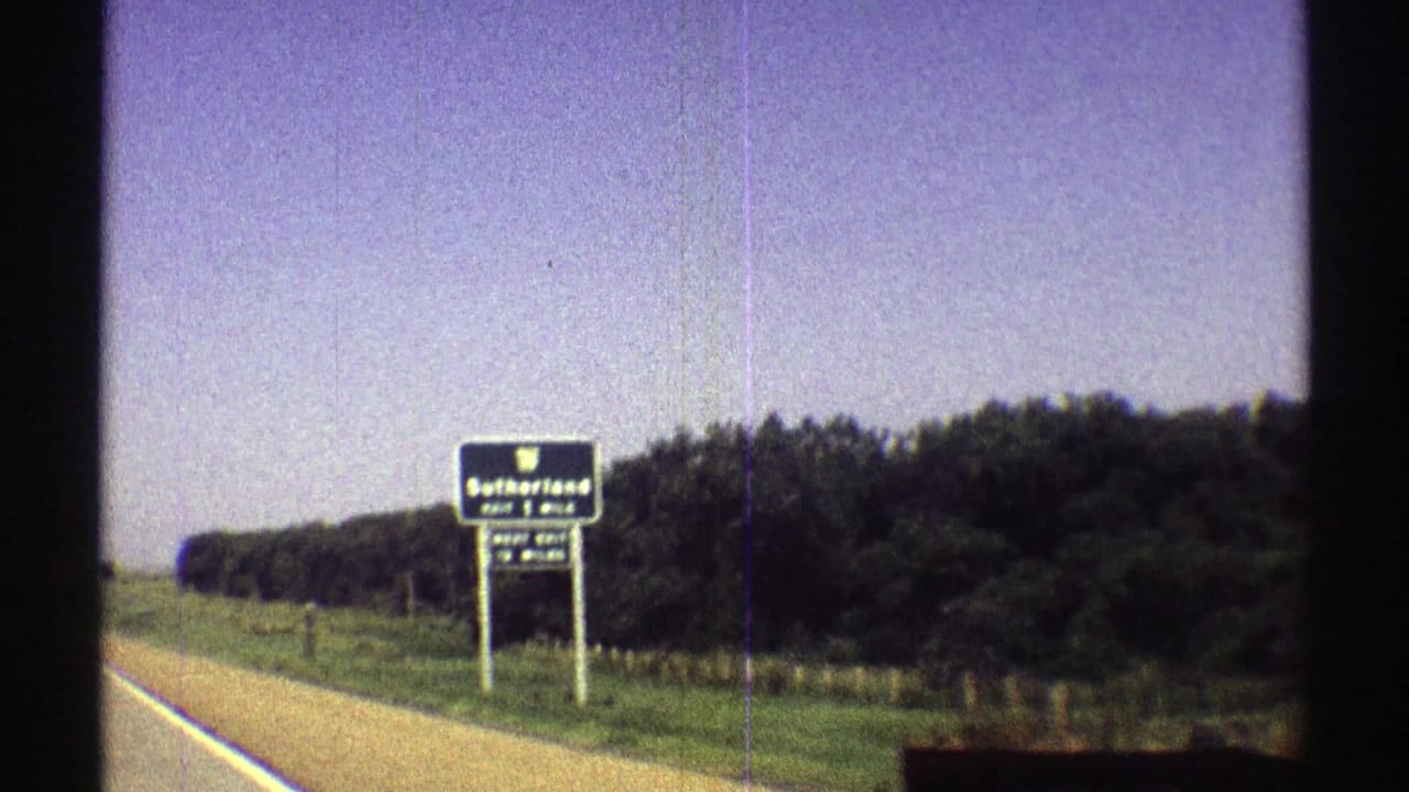 Blue highway sign for Sutherland Exit 1 Mile beside a rural road and trees.