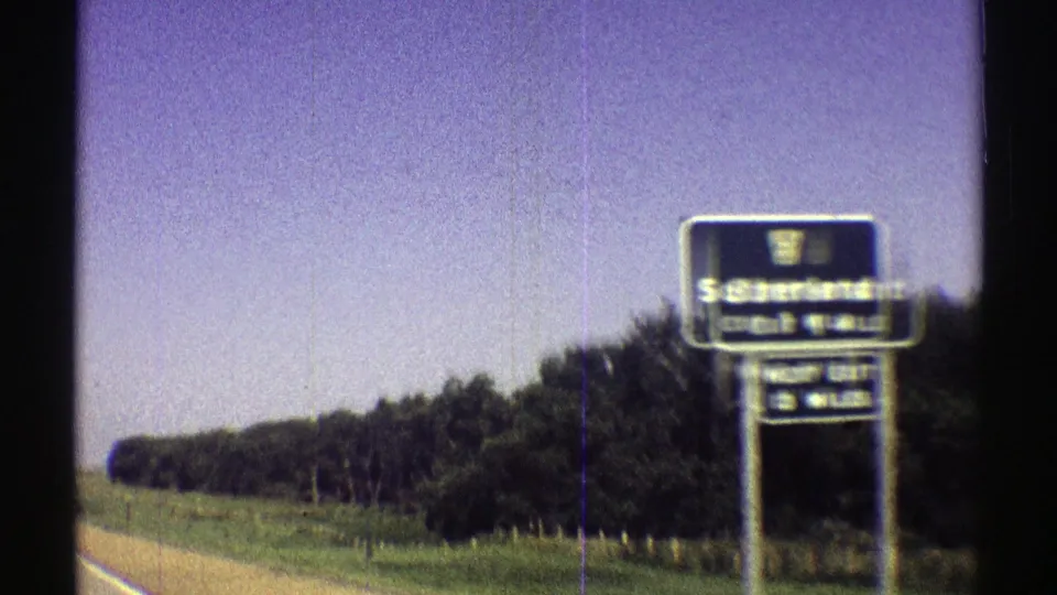 1969 Nebraska 8mm frame with the Sutherland exit sign filling more of the view as the car passes roadside trees.
