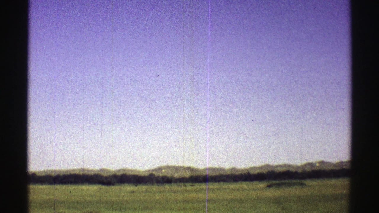 1969 Nebraska 8mm frame showing open green prairie, a distant tree line, and low hills beneath a pale purple sky.