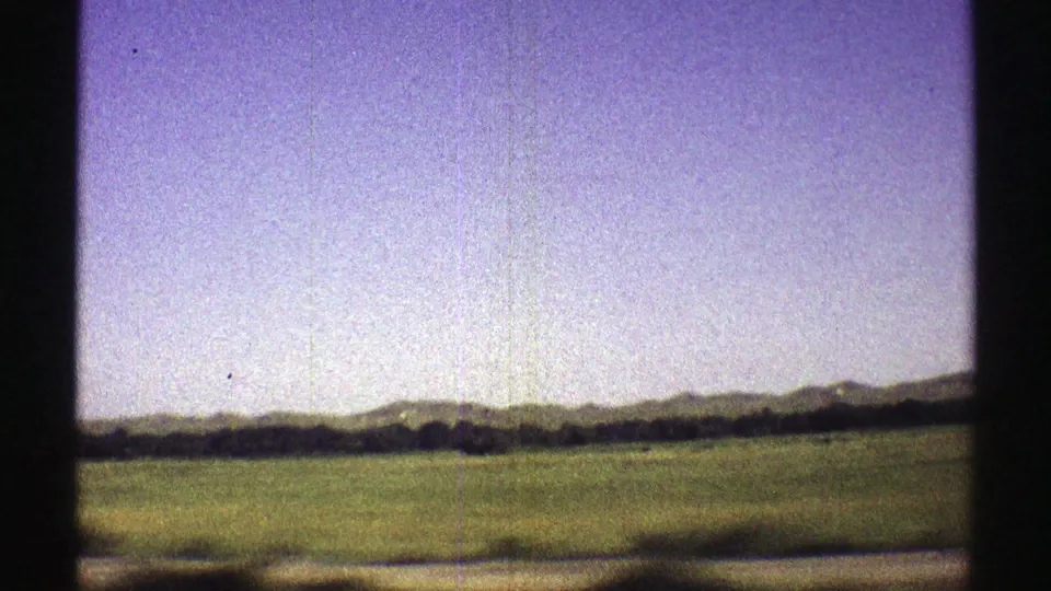 1969 Nebraska 8mm frame showing a green roadside field, dark horizon line, and low hills under a washed sky.