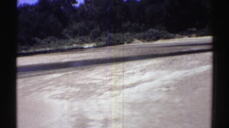 Nebraska stream bank with a broad pale sand bar, shallow water, and thickets of trees in 1969 home-movie footage.
