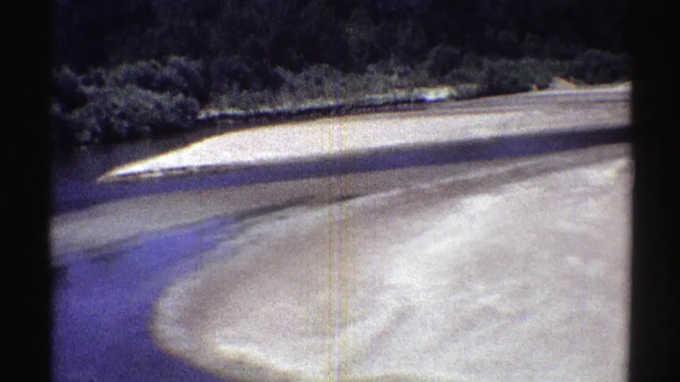 Curving sand bar beside a slow Nebraska stream, with dark trees and brush forming the far bank in 1969 8mm footage.