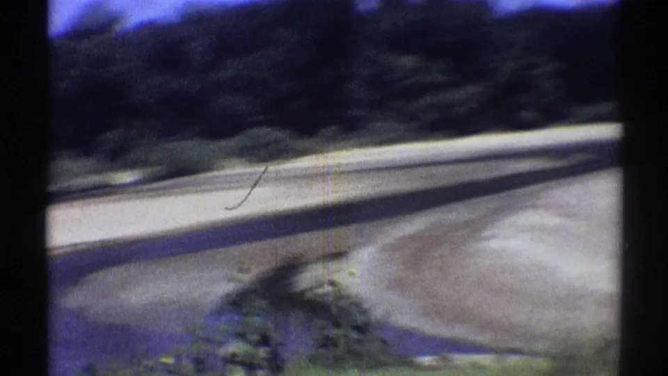 A slow stream channel cuts past pale sand and a dark wooded bank in archival 1969 Nebraska 8mm footage.