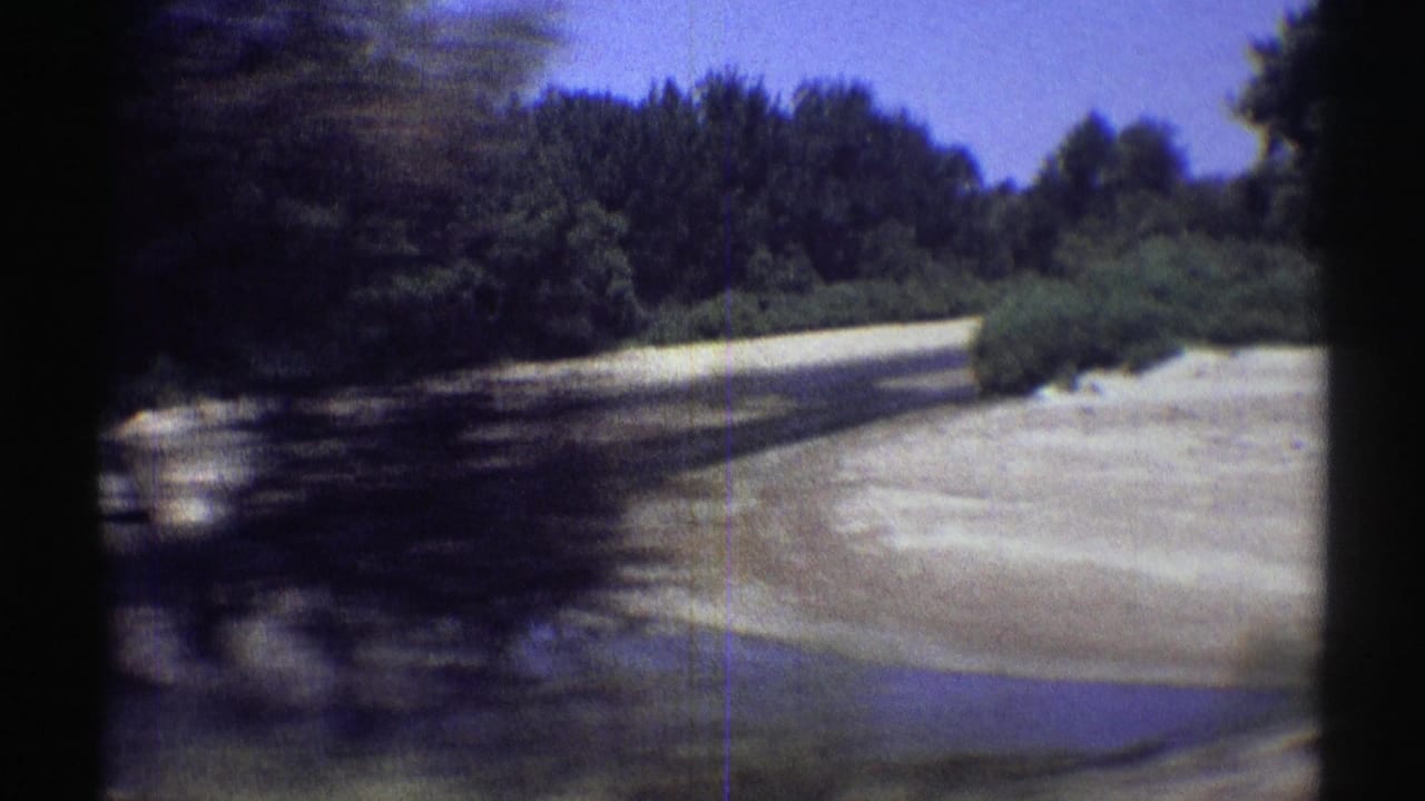 1969 Nebraska 8mm frame from a moving car showing trees, a pale bank, a small water channel, and a roadside curve.