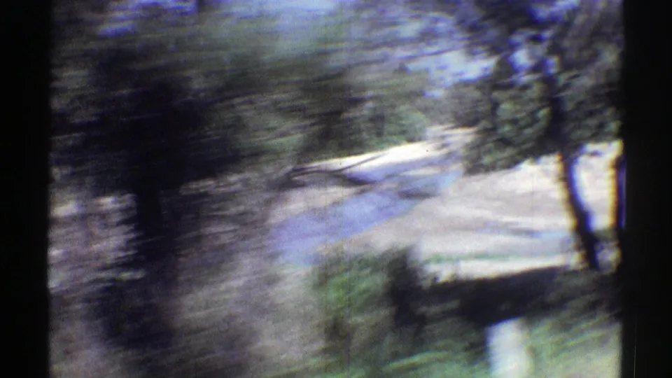 Moving 8mm view in rural Nebraska with blurred trees, a light riverbank, brush, and dark vertical film-frame borders.