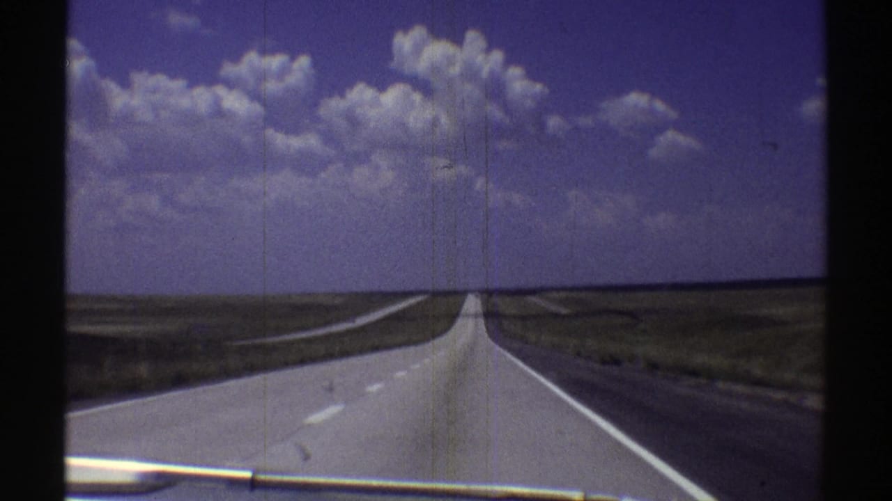 1969 Nebraska 8mm view from a moving car toward a straight grey highway beneath cumulus clouds.
