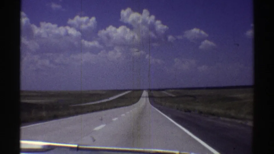 1969 Nebraska 8mm view from a moving car toward a straight grey highway beneath cumulus clouds.