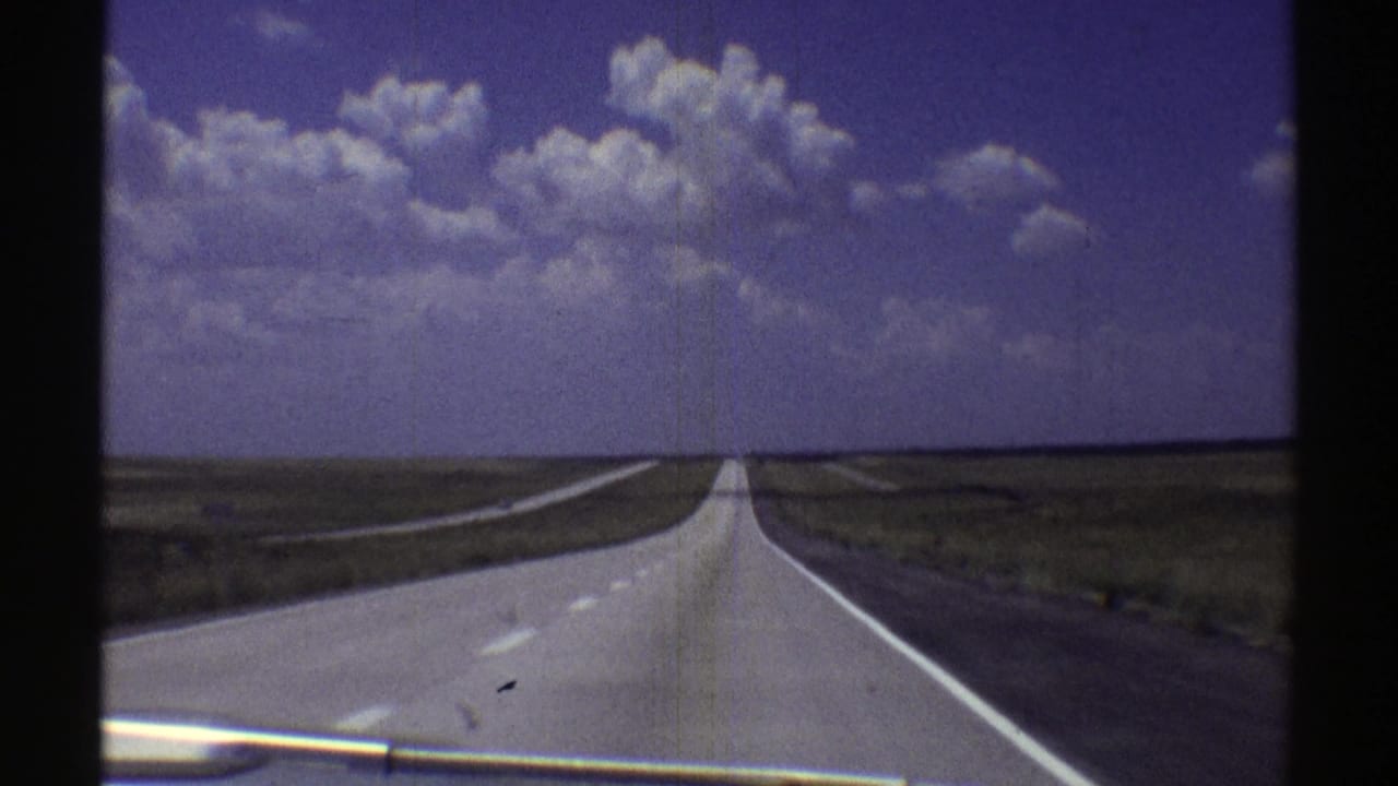 Forward-facing car-window view of a straight Nebraska highway in 1969 with open plains and a blue, clouded sky.