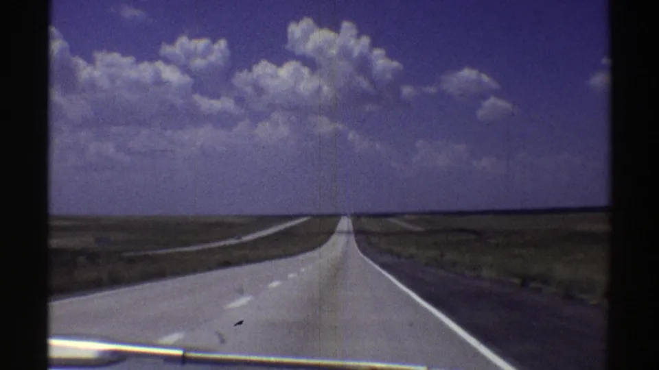 Forward-facing car-window view of a straight Nebraska highway in 1969 with open plains and a blue, clouded sky.