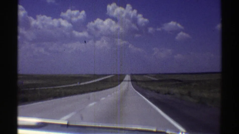 Vintage 8mm driving shot along a straight Nebraska highway, with dashboard trim visible at the bottom of the frame.
