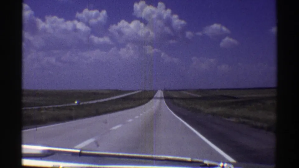 Late-1960s home-movie frame of a Nebraska highway vanishing toward the horizon under bright cumulus clouds.
