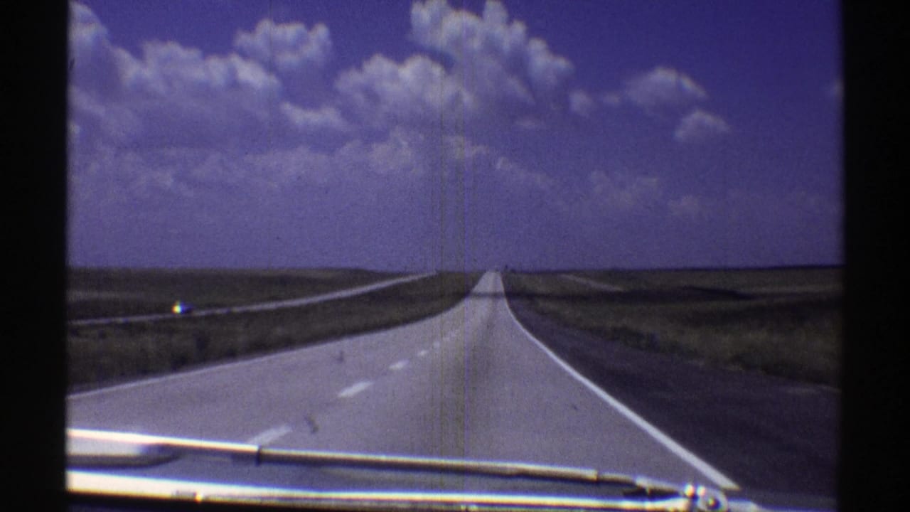 1969 Nebraska 8mm frame showing a straight pale asphalt highway across open plains beneath a blue sky with white cumulus clouds, with a windshield wiper visible at the bottom.