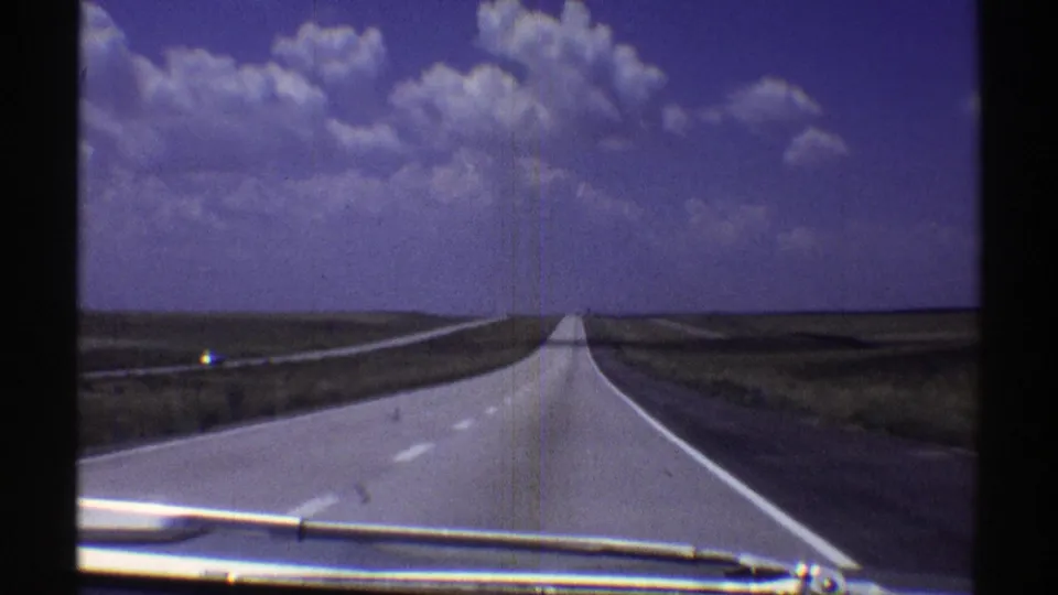 1969 Nebraska 8mm frame showing a straight pale asphalt highway across open plains beneath a blue sky with white cumulus clouds, with a windshield wiper visible at the bottom.