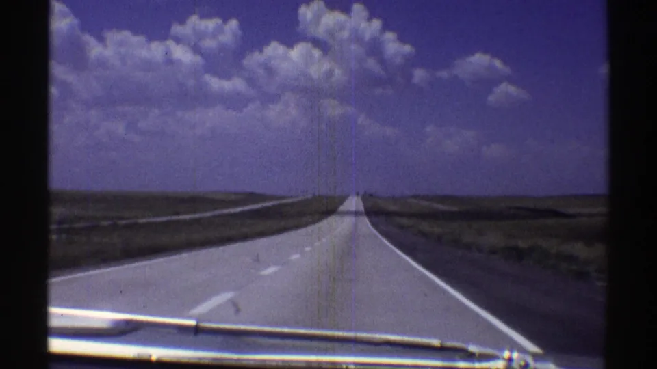 Forward-facing 1969 Nebraska road view from inside a moving car, with the light grey highway narrowing toward the horizon under scattered white clouds.