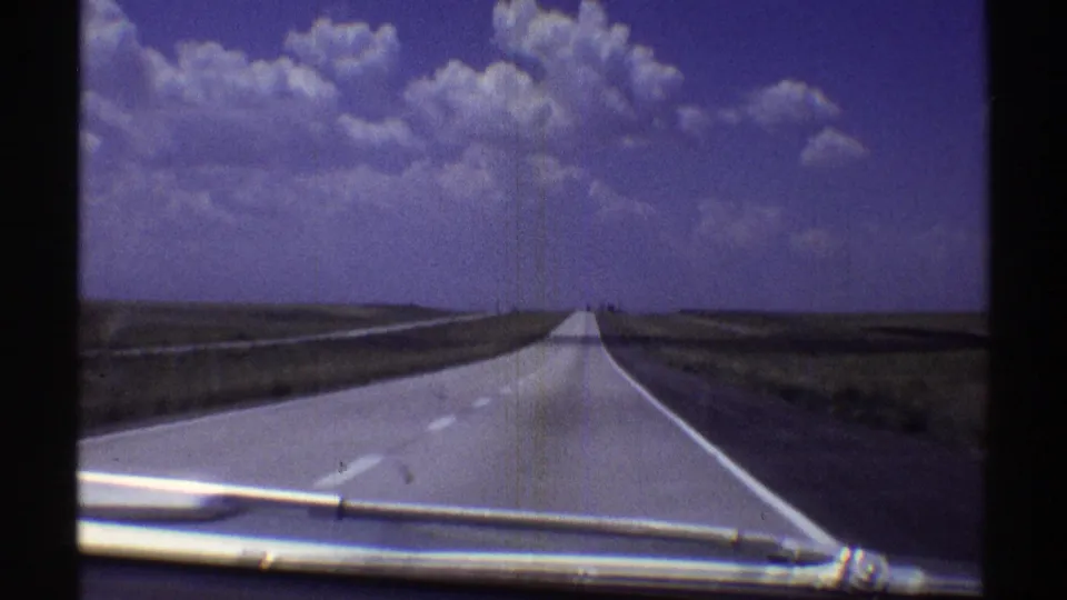 Vintage 8mm view of a straight Nebraska highway across flat rural terrain, with soft film grain, blue-purple color shift, and windshield hardware at the lower frame edge.