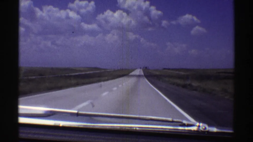 Final 1969 Nebraska frame looking forward through a windshield at a straight rural highway, low grassland edges, and a bright sky filled with cumulus clouds.