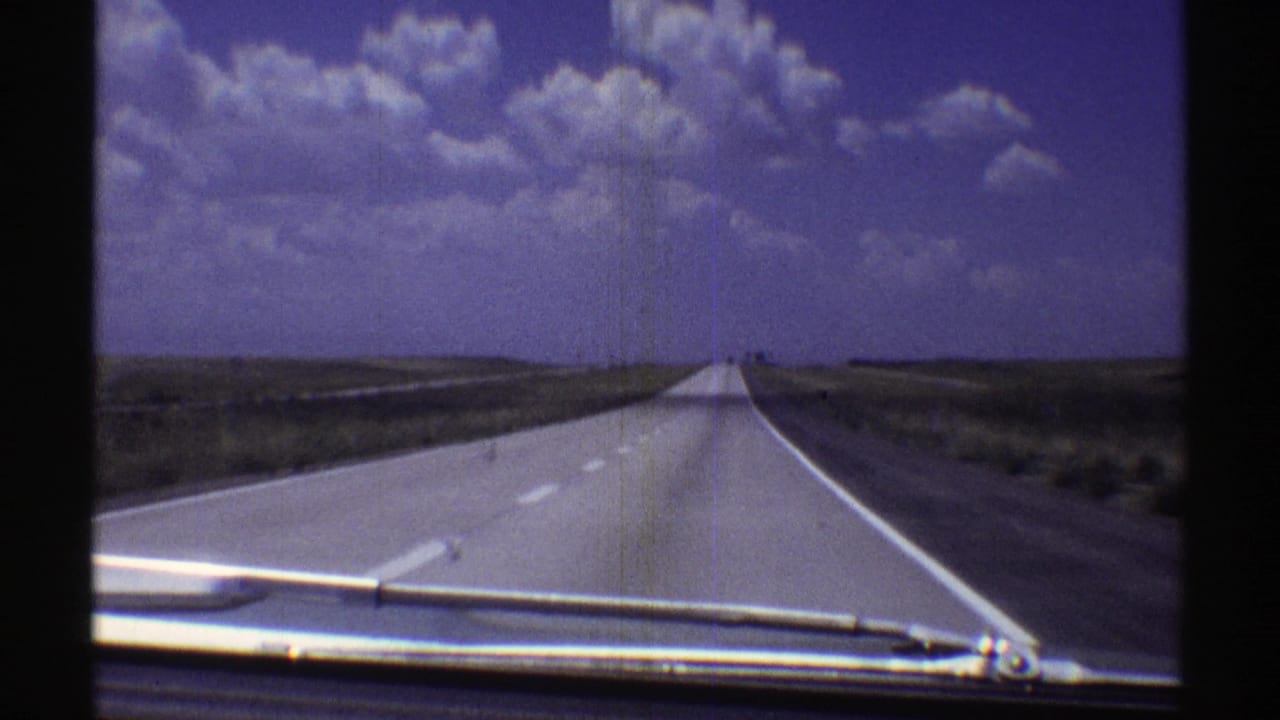 1969 Nebraska 8mm view down a straight rural highway, with car hood and windshield wiper visible under large clouds.