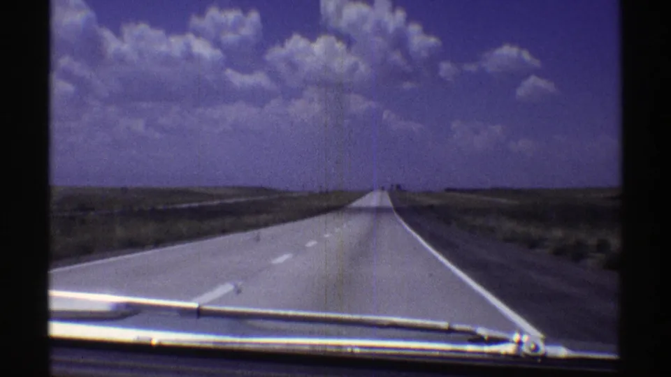 1969 Nebraska 8mm view down a straight rural highway, with car hood and windshield wiper visible under large clouds.