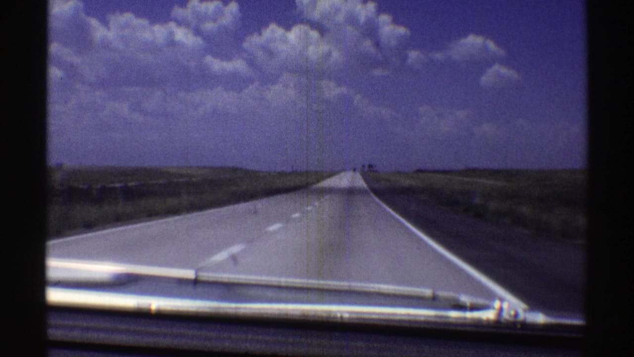 Forward-facing 1969 Nebraska road view with a pale highway narrowing toward the horizon and clouds above the plains.