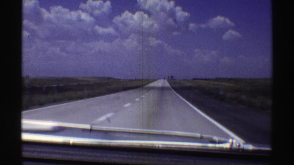 Forward-facing 1969 Nebraska road view with a pale highway narrowing toward the horizon and clouds above the plains.