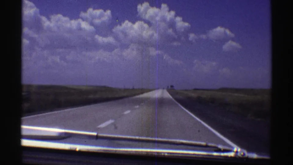 1969 Nebraska home-movie frame showing a rural highway, low plains, and a vivid clouded sky from a car viewpoint.